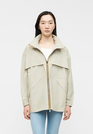 Joven con cabello largo y oscuro, lleva una chaqueta beige clara con cremallera y jeans azules claros, de pie contra un fondo blanco liso.