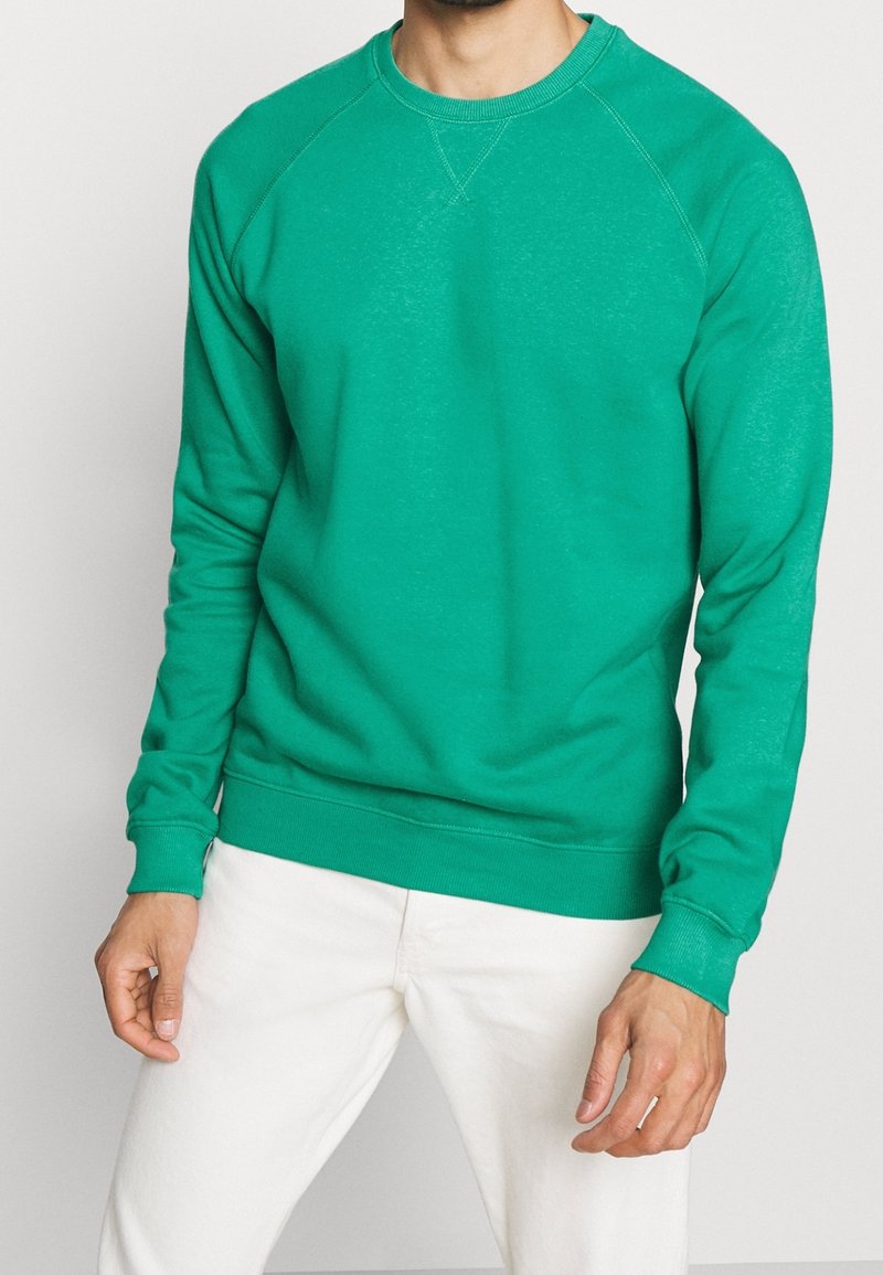 Sudadera de algodón verde con cuello redondo, mangas raglán largas, puños y bajo de canalé, exhibida sobre pantalones blancos. Sin patrones ni acentos.