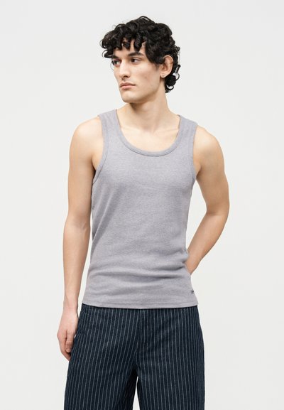 Νέος άνδρας με κατσαρά μαλλιά που φοράει αμάνικη ανοιχτογκρι μπλούζα και σκουρόχρωμο παντελόνι με λεπτές ρίγες, στέκεται με το ένα χέρι πίσω από την πλάτη του.