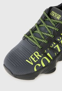 Sapatilha atlética cinza com parte superior texturizada, detalhes pretos, atacadores verde néon e uma marcação amarela oversized na lateral. Design da sola curvada.