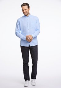 Lichtblauw overhemd met lange mouwen en knopen, voorzien van een borstzak, gedragen met donkere broek en witte sneakers. Soepele textuur, relaxte pasvorm.