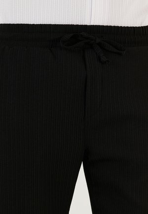 Črne rebraste trenirke z elastičnim pasom in detajlom vrvice. Tekstura je mehka z navpičnimi črtami, ki ustvarjajo subtilen vzorec.