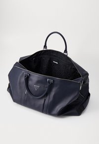 Sac de voyage en cuir bleu marine avec des poignées supérieures doubles, fermeture éclair et doublure intérieure à motifs. Comprend une plaque logo sur le devant et des poches latérales.