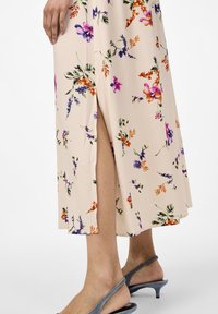 Robe midi florale en beige clair, ornée de fleurs multicolores, avec une fente sur le côté et une texture fluide, assortie à des sandales à talons grises.