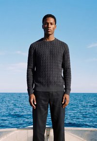 Jeune homme portant un pull torsadé foncé et un pantalon noir, debout sur un bateau avec la mer bleue et le ciel en arrière-plan.