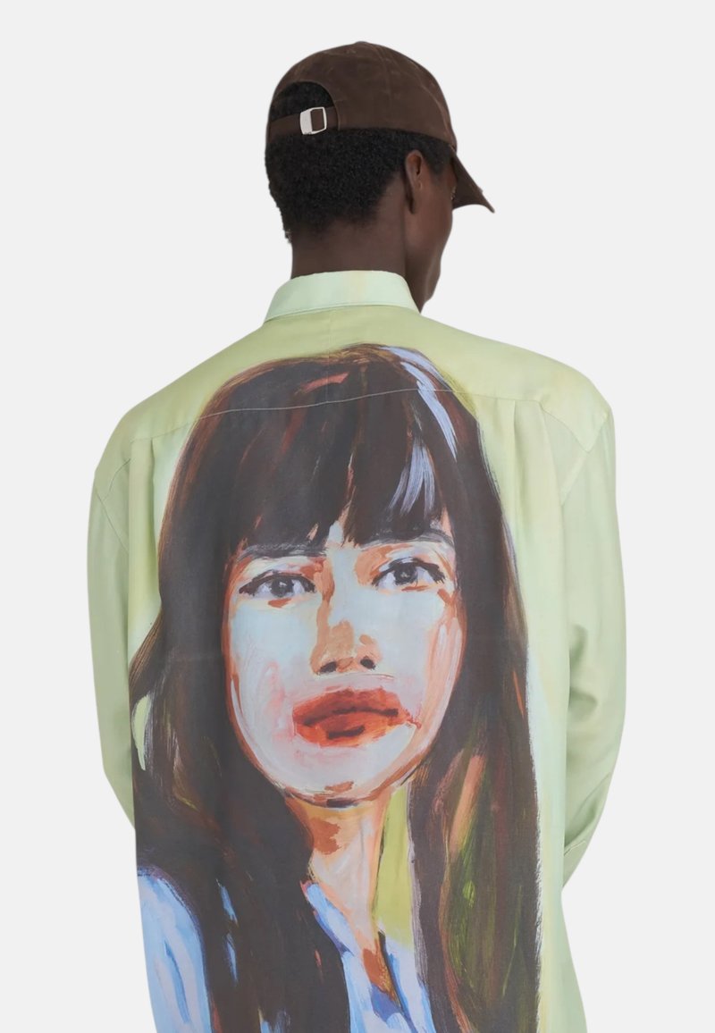 Chemise oversize verte clair à manches longues, ornée d'un portrait peint au dos. Tissu lisse, coupe décontractée et œuvre d'art du visage détaillée.