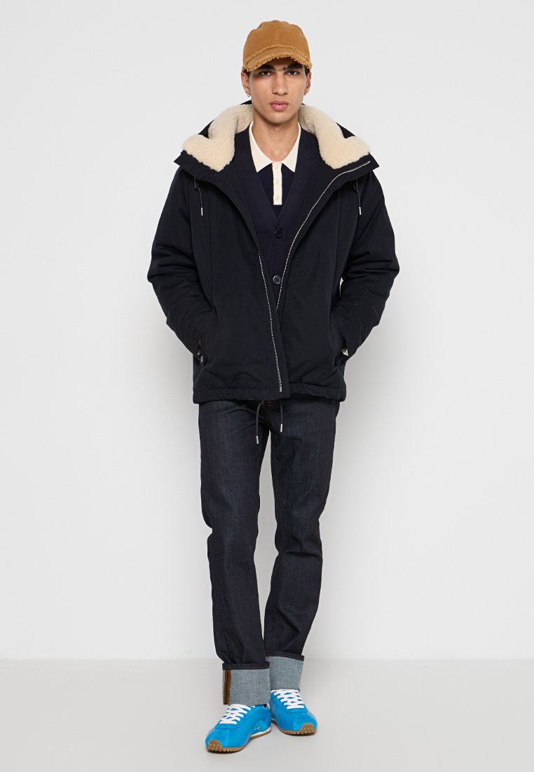 Marine parka met een faux-fur gevoerd capuchon, knoopsluitingen, gelaagd over een marineblauwe trui en een lichtkleurig overhemd. Donkere jeans en blauwe sneakers.