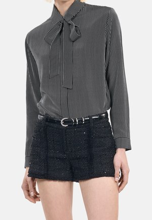 Kvinde iført en sort og hvid vertikalt stribet bluse med nakkesløjfe og sorte teksturerede shorts med sort bælte.