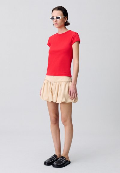Maglietta rossa aderente a maniche corte, gonna beige a volant, scarpe nere slip-on e occhiali da sole bianchi. Design semplice e moderno con un vivace contrasto di colori.