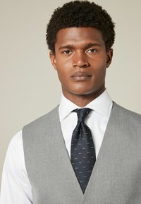 Gray vest over a white shirt, paired with a navy tie featuring small patterns. Close-up of a person's upper body. Textured fabric detail visible.