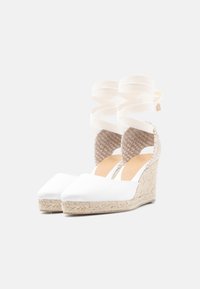 Λευκά παπούτσια εσπαντρίγιες με πλεκτές φτέρνες και κορδέλες που δένονται γύρω από τον αστράγαλο, σε απλό λευκό φόντο.