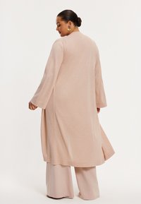 Langer, hellrosa Cardigan mit einem dezenten Schimmer, mit weiten Ärmeln und seitlichen Schlitzen, kombiniert mit hellbeigen, weitem Hosenbeinen.