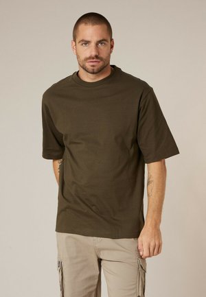 Homme aux cheveux courts et à la barbe, portant un T-shirt vert olive foncé et un pantalon cargo beige, debout devant un fond beige uni.