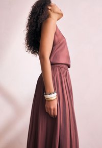 Mauvefarbenes, ärmelloses Oberteil mit einem fließenden, plissierten Rock, der eine glatte Textur zeigt. Accessoires umfassen gestapelte Armreifen aus verschiedenen Materialien.