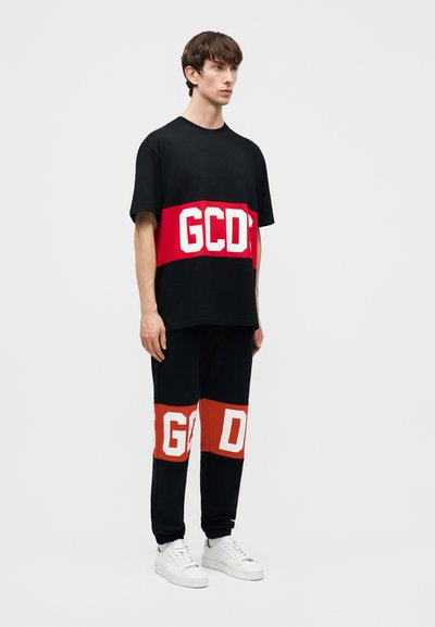 Ung mand iført sort outfit med røde bånd og hvidt "GCDS"-tryk, iført hvide sneakers, mod en ensartet lys baggrund.