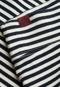 Gestreepte stof met afwisselend marineblauwe en crèmekleurige strepen, met een geweven rode label waarop "Vintage Essentials" in een textuurontwerp staat.