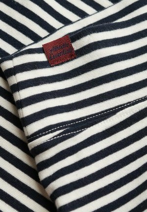 Tkanina w paski z naprzemiennymi niebiesko-granatowymi i kremowymi paskami, z tkanym czerwoną metką z napisem "Vintage Essentials" w teksturowanym wzorze.