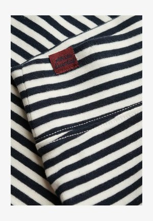 Tela a rayas con franjas alternadas en azul marino y crema, con una etiqueta tejida en rojo que muestra "Vintage Essentials" en un diseño texturizado.