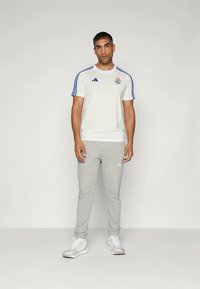 Camiseta deportiva blanca con Rayas azules y logotipo, combinada con pantalones de chándal grises con rayas laterales, y zapatillas blancas.