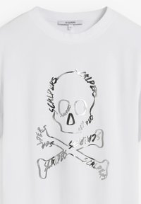 Bílý bavlněný tričko s lesklým stříbrným designem lebky a zkřížených kostí, na kterém se opakuje text "Scalpers" v graffiti stylu.