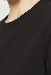 Primer plano del cuello y el hombro de una persona que lleva una camiseta negra de cuello redondo sobre un fondo claro.