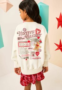 Cream-colored oversized sweatshirt featuring festive gingerbread baking graphics, red accents, and a pink sprinkle pattern. Red sequined skirt below.