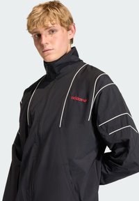 Black athletic jacket with high collar, white stripes on sleeves, and red "adidas" logo. Lightweight fabric with a smooth texture.