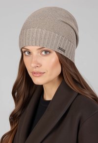 Beige geribbelde gebreide muts met een omgeslagen rand en een metalen merkteken. Zachte textuur, ontworpen om prettig en strak op het hoofd te passen.