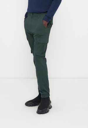 Muž oblečený v tmavozelených cargo nohaviciach s elastickými manžetami, čiernych slip-on hrubých teniskách a tmavomodrom tričku s dlhým rukávom, ruky v vreckách.