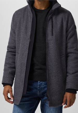 Man draagt een donkergrijs jack met rits over een zwarte trui en een blauwe spijkerbroek, staande tegen een effen lichte achtergrond.