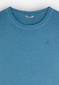 Camiseta de color azul claro hecha de un tejido suave. Presenta un cuello redondo y un pequeño logo de calavera en la parte izquierda del pecho. Textura lisa.