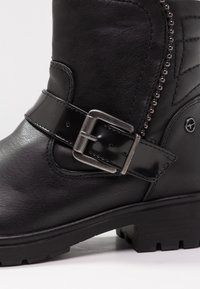 Bottes noires en cuir avec une sangle brillante et une boucle en argent, finition texturée et semelle robuste avec des crampons proéminents pour une bonne traction.