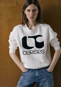 Sudadera blanca con un gran logotipo gráfico negro y el texto "cerises", combinada con unos jeans azules desgastados. Tejido suave y corte relajado.