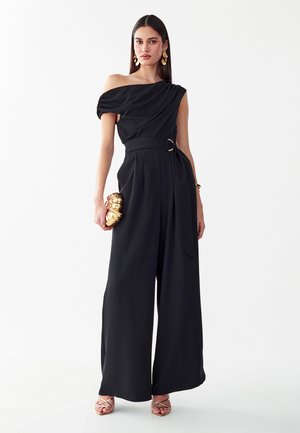 Femme portant une combinaison noire asymétrique avec une ceinture, des boucles d'oreilles dorées, tenant une pochette dorée et portant des talons dorés à brides.