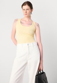 Yellow ribbed tank top with a scoop neckline, paired with high-waisted white trousers. Holding a black quilted handbag.