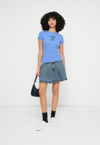 Tricou albastru cu dungi, cu mâneci scurte și imprimeu negru, asortat cu o fustă din denim. Modelul poartă o geantă mică neagră și adidași albi.