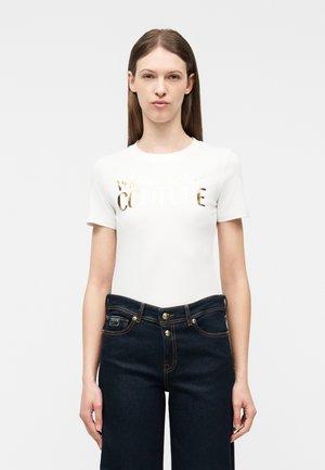 Γυναίκα με ίσια καστανά μαλλιά φοράει λευκό κοντομάνικο μπλουζάκι με χρυσό κείμενο και σκουρόχρωμο μπλε τζιν ψηλόμεσο, στέκεται μπροστά σε απλό φόντο.