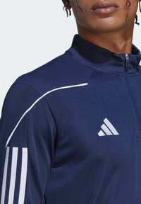 Giacca zip-up blu navy con spalle testurizzate, dettagli con strisce bianche e logo bianco sul lato sinistro del petto. Presenta un colletto alto.