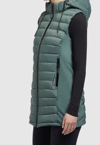 Bez rękawów zielona pikowana kamizelka z przednim zamkiem błyskawicznym i kapturem, noszona na czarnej bluzie z długim rękawem, pokazana z boku na stojącej osobie.