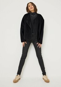 Czarna polarowa kurtka z opadającym przodem, połączona z czarnymi obcisłymi dżinsami i beżowymi sneakersami. Stylizacja zawiera subtelną biżuterię.