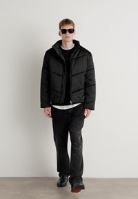 Black puffer jacket with quilted design, worn over a black shirt and white t-shirt. Black pants and shoes with red accents complete the look.