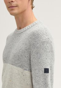 Hellgrauer Strickpullover mit strukturiertem Finish und cremefarbener Streifen. Verfügt über einen Rundhalsausschnitt und ein schwarzes Logo-Patch auf dem linken Ärmel.