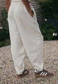 Baltos medvilninės sportinės kelnės su šoninėmis kišenėmis, elastine juosmens dalimi ir siaurėjančiomis kojomis, dėvimos kartu su rudais ir kreminiais kedais ant žvyro paviršiaus.