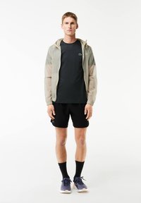 Giacca leggera e trasparente con cappuccio, abbinata a una maglietta scura e pantaloni corti neri. Il modello indossa scarpe da ginnastica viola e calzini neri.