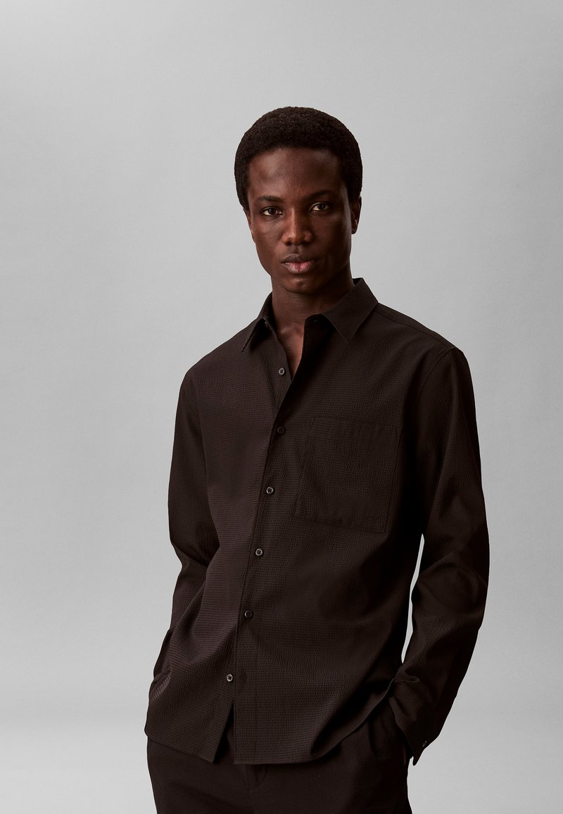 Jeune homme à la peau foncée portant une chemise noire texturée à boutons, debout les mains dans les poches devant un fond gris uni.