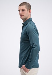 Ilgomis rankovėmis polo marškinėliai žalsvos spalvos audinyje, turintys apykaklę, liekną fasoną, lygų paviršių ir subtilų logotipą kairėje krūtinės pusėje.