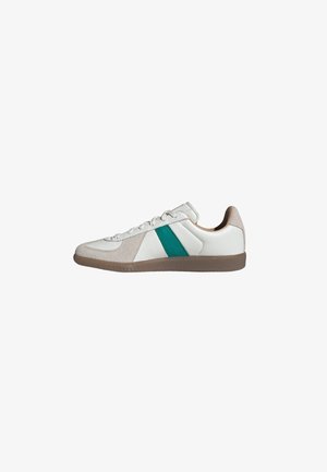 Zapatilla deportiva de piel blanca con detalles en ante beige, raya lateral verde y suela de goma marrón, mostrada de perfil.