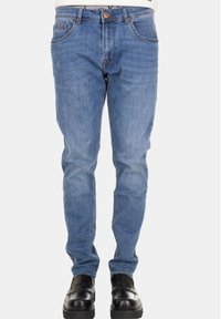 Uomo che indossa jeans slim-fit azzurri e scarpe slip-on in pelle nera, in piedi su uno sfondo bianco neutro.