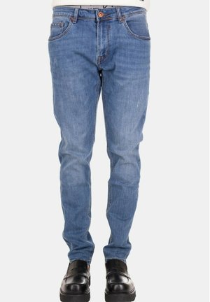 Uomo che indossa jeans slim-fit azzurri e scarpe slip-on in pelle nera, in piedi su uno sfondo bianco neutro.