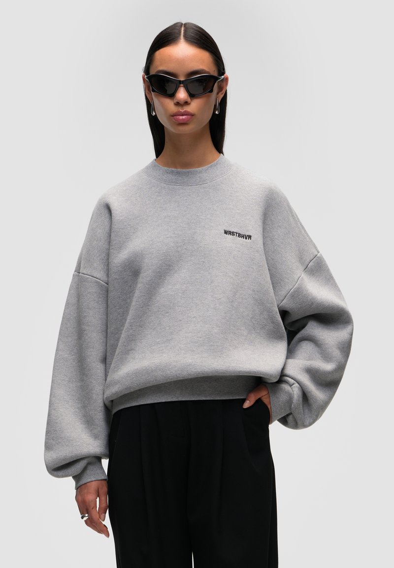 Felpa grigia oversize con spalle scese e polsini a costine. Presenta un piccolo logo nero sulla parte anteriore. Abbinata a pantaloni neri.